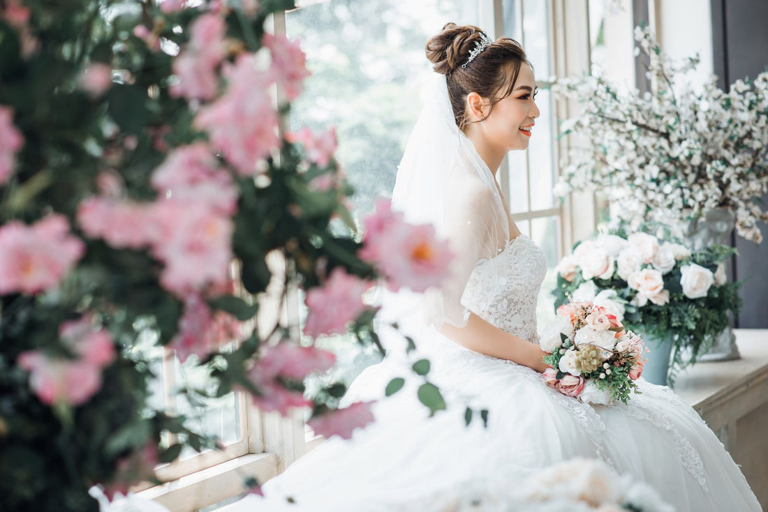 Bride in a white gown holding flowers by window light; editorial inspiration photo, not a Rosy Flower Wall product or client event.