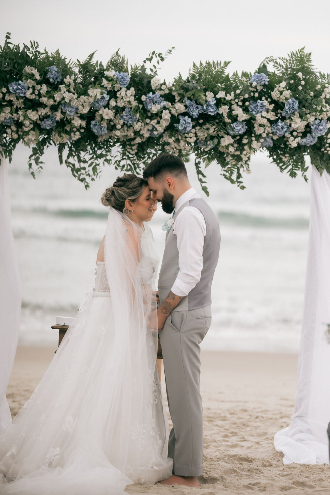 Couple at a beach wedding ceremony beneath a floral arch beside the ocean