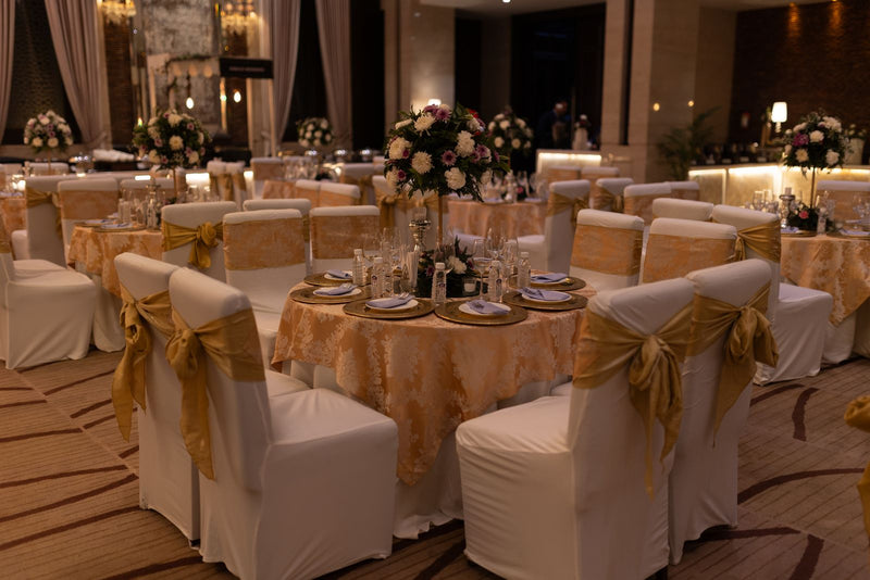 Large indoor wedding reception hall with round tables, floral arrangements, chandeliers, and clear aisles