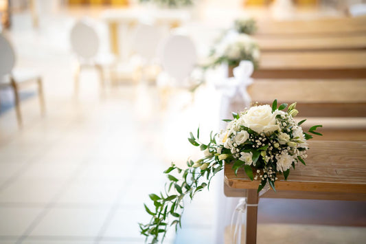 White roses and greenery arranged along wooden benches in an indoor wedding aisle
